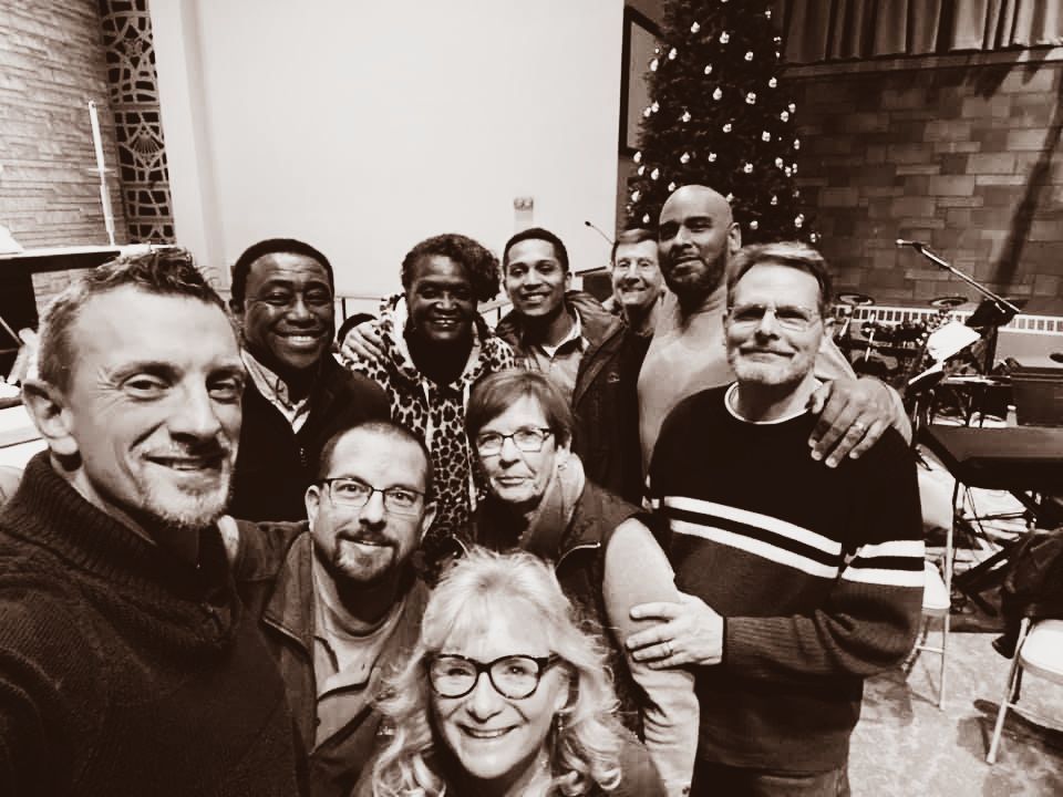 Group selfie of diverse people smiling in a room, near a Christmas tree.