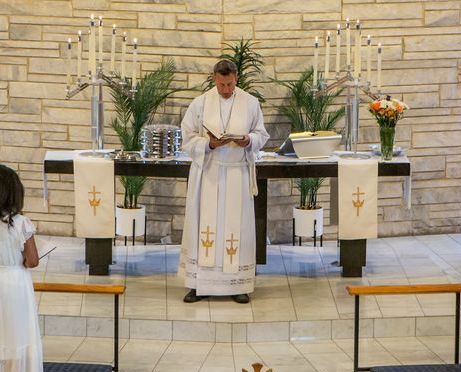 A priest in white robes reads from a book at an altar. A child stands nearby holding a candle.