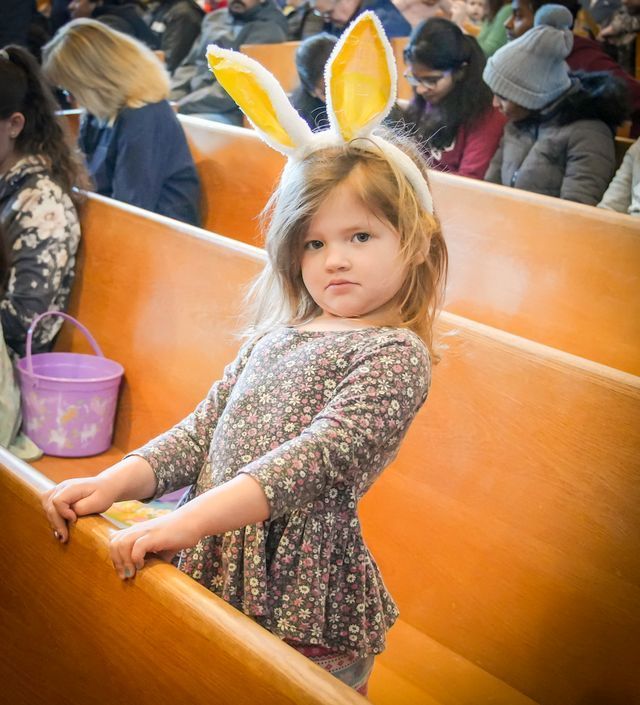 Young girl in floral dress and bunny ears stands in church pew, looking unimpressed.