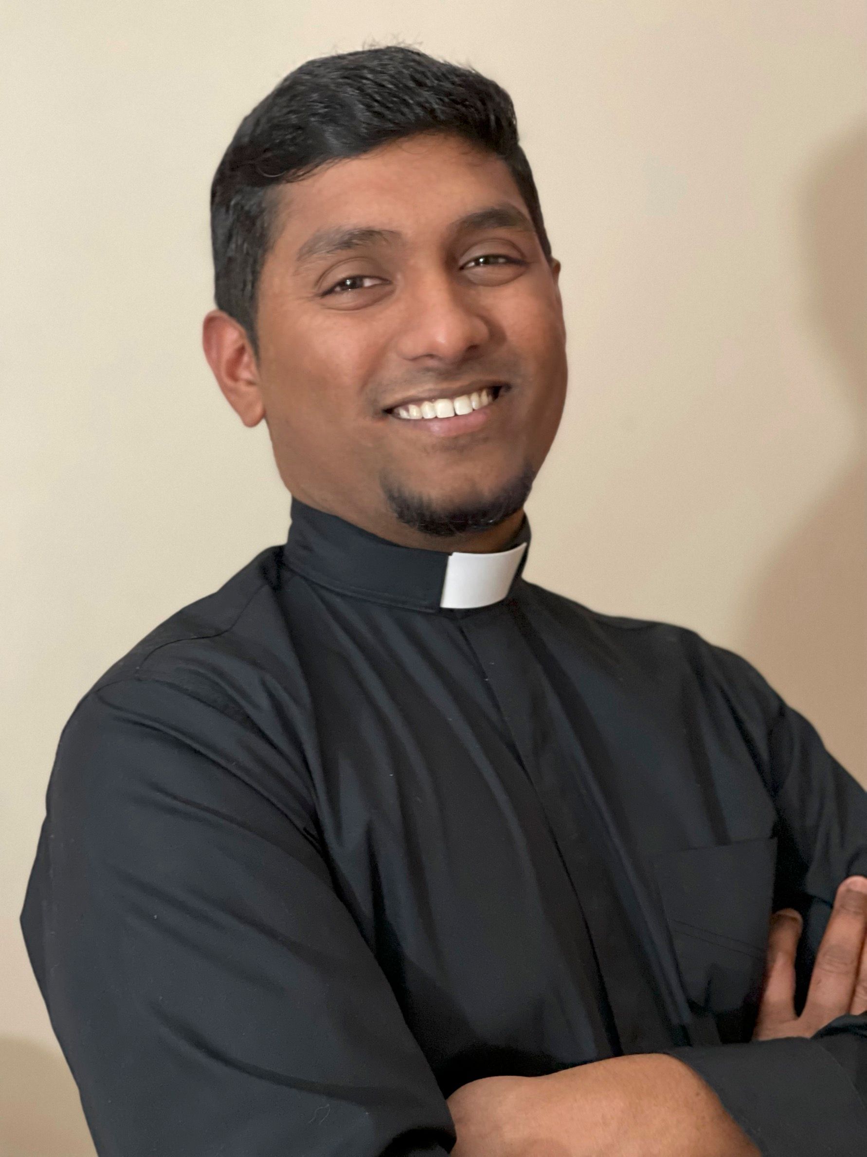 The Vicar in a black robe with a white collar is smiling with his arms crossed