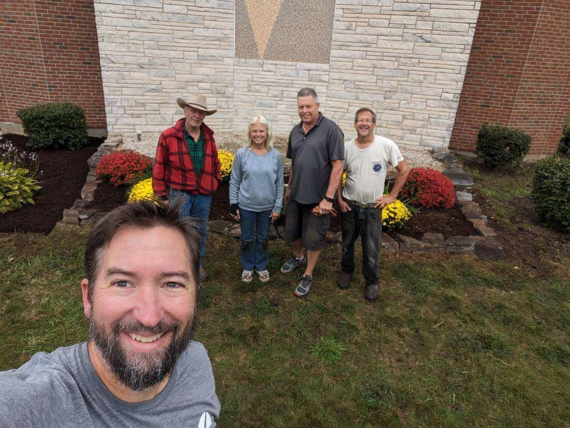 Group of five people posing in front of a building, taking a selfie. Green grass, flowers, and brick.