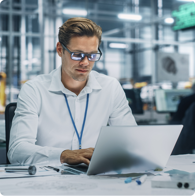 Man in glasses typing on a laptop, in a bright industrial setting.