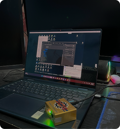 Laptop displaying software, connected to a gold box on a desk. Other screens and a mouse are visible.