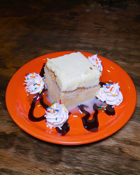 Square slice of white-frosted cake on an orange plate, drizzled with chocolate and whipped cream rosettes.
