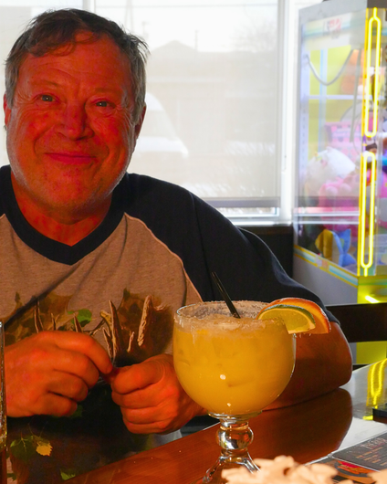 Man sitting at a table with a large margarita in a neon-lit bar, smiling.