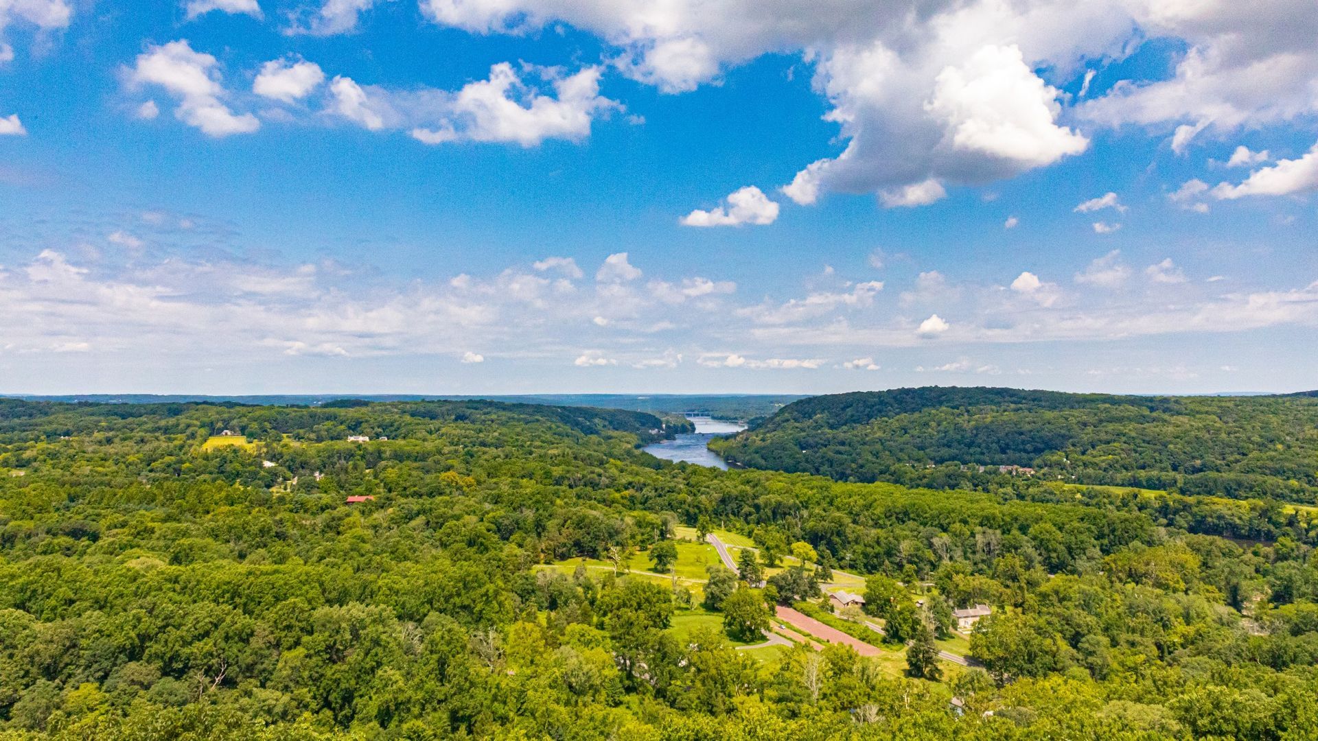 Wide aerial view of lush green forest and rolling hills under a blue sky with clouds and a distant river in Bucks County, PA
