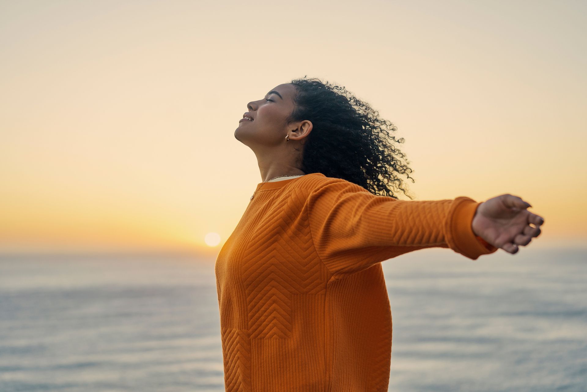 A woman in an orange sweater is standing on the beach with her arms outstretched.
