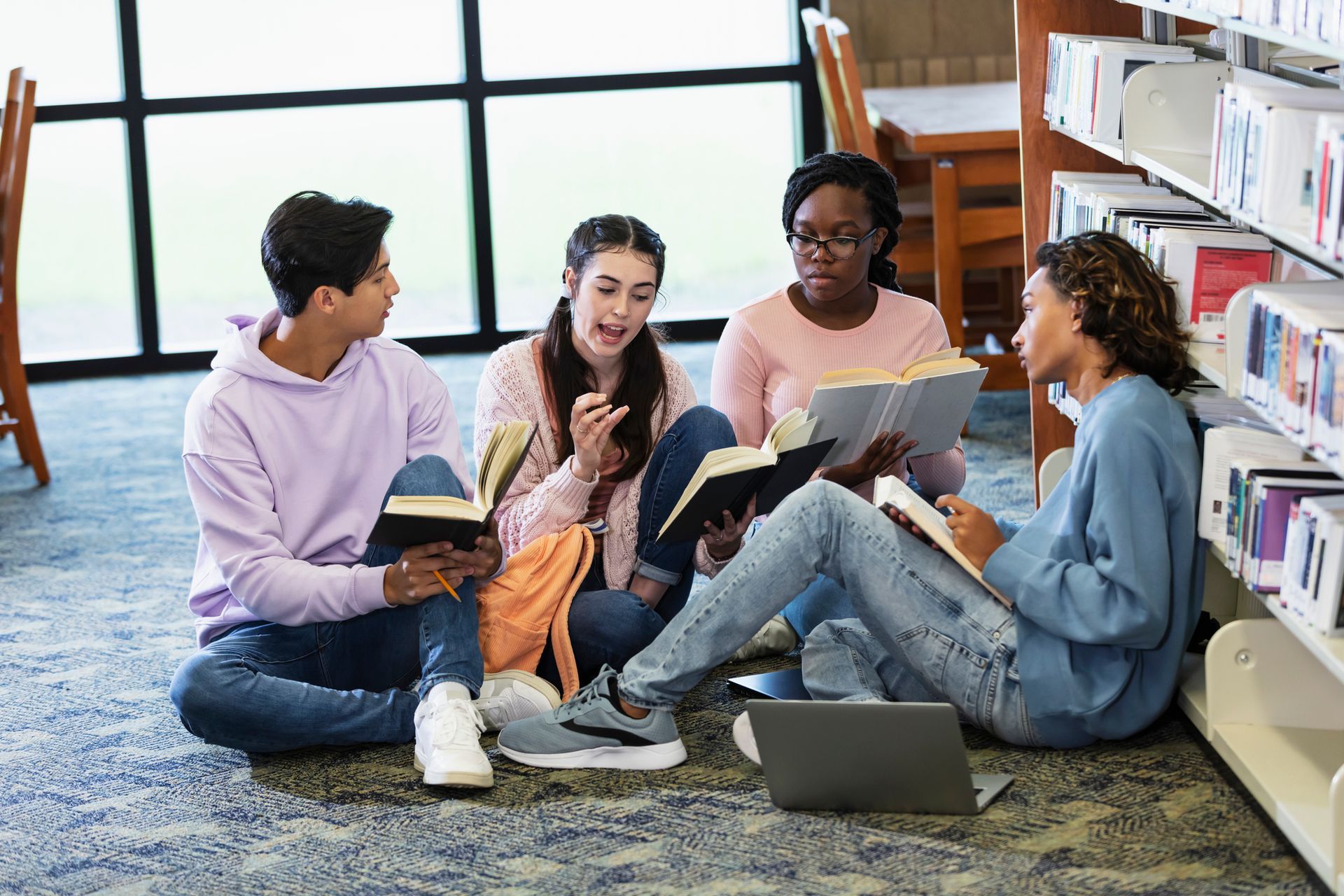 A group of young people are sitting on the floor in a library reading books.