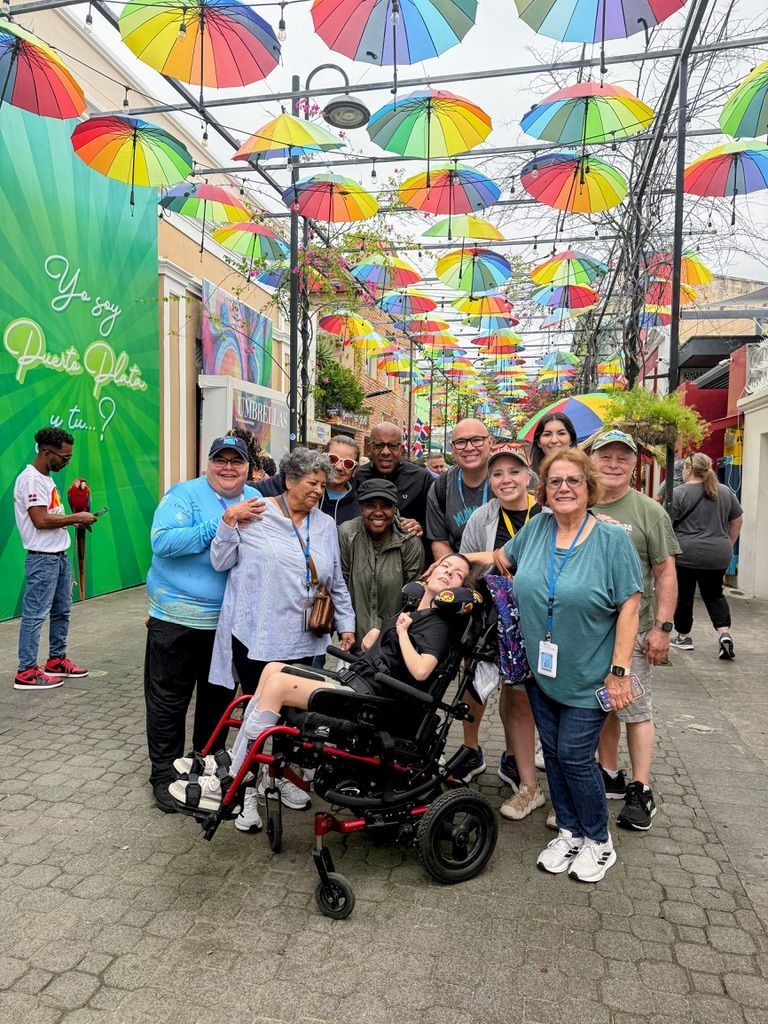 Group gathered under rainbow umbrellas in a lively outdoor walkway, smiling around a wheelchair user.