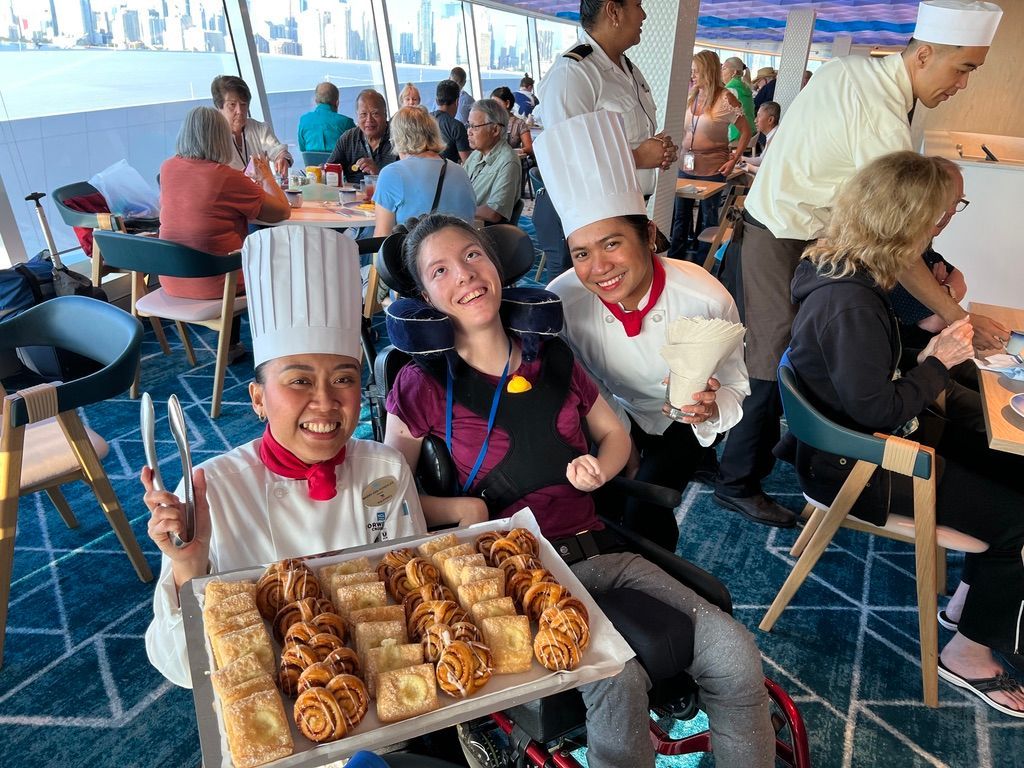 Three smiling chefs holding a tray of pastries on a boat deck with other passengers in the background