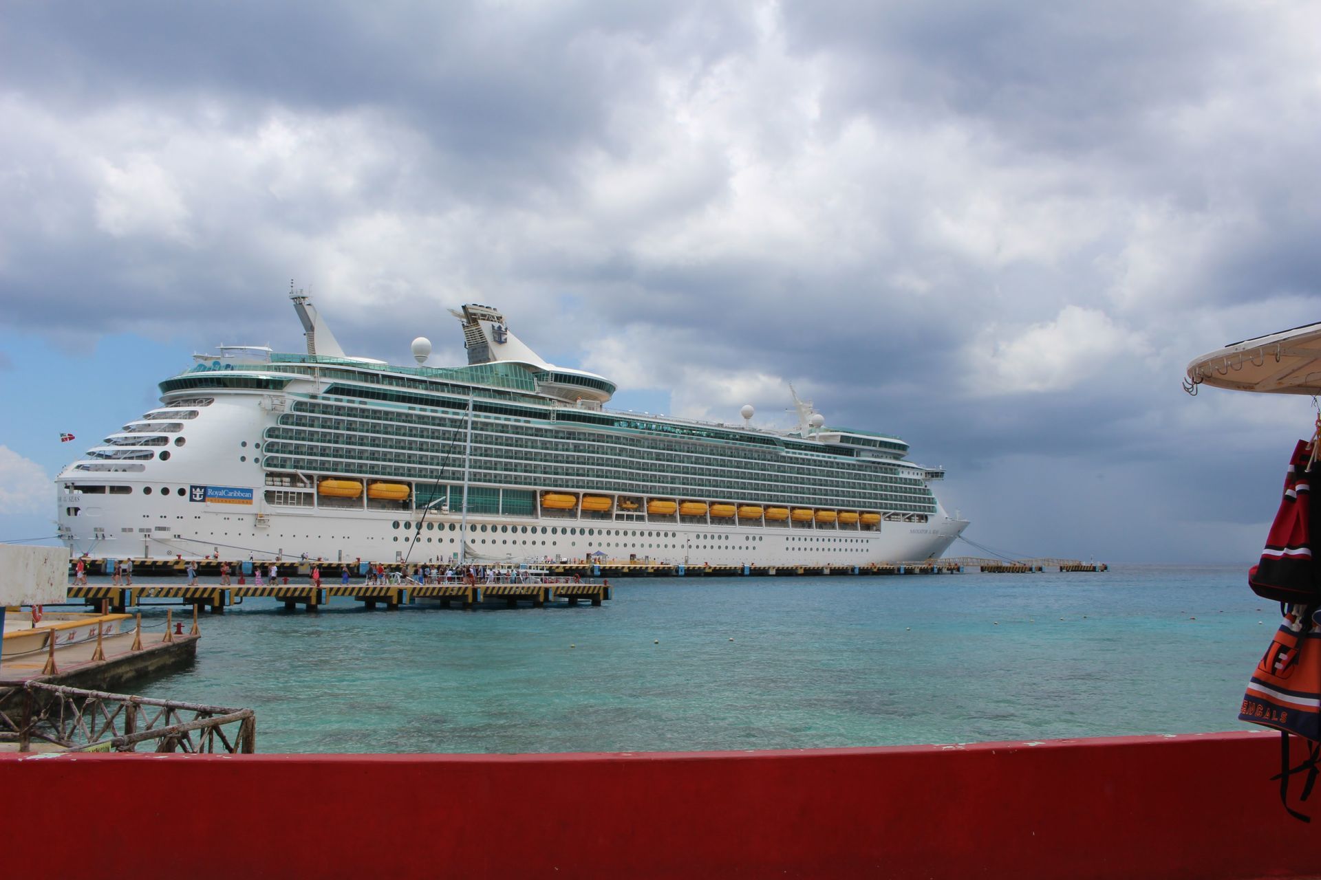 Large white cruise ship docked in turquoise water under cloudy skies, seen from a red pier railing