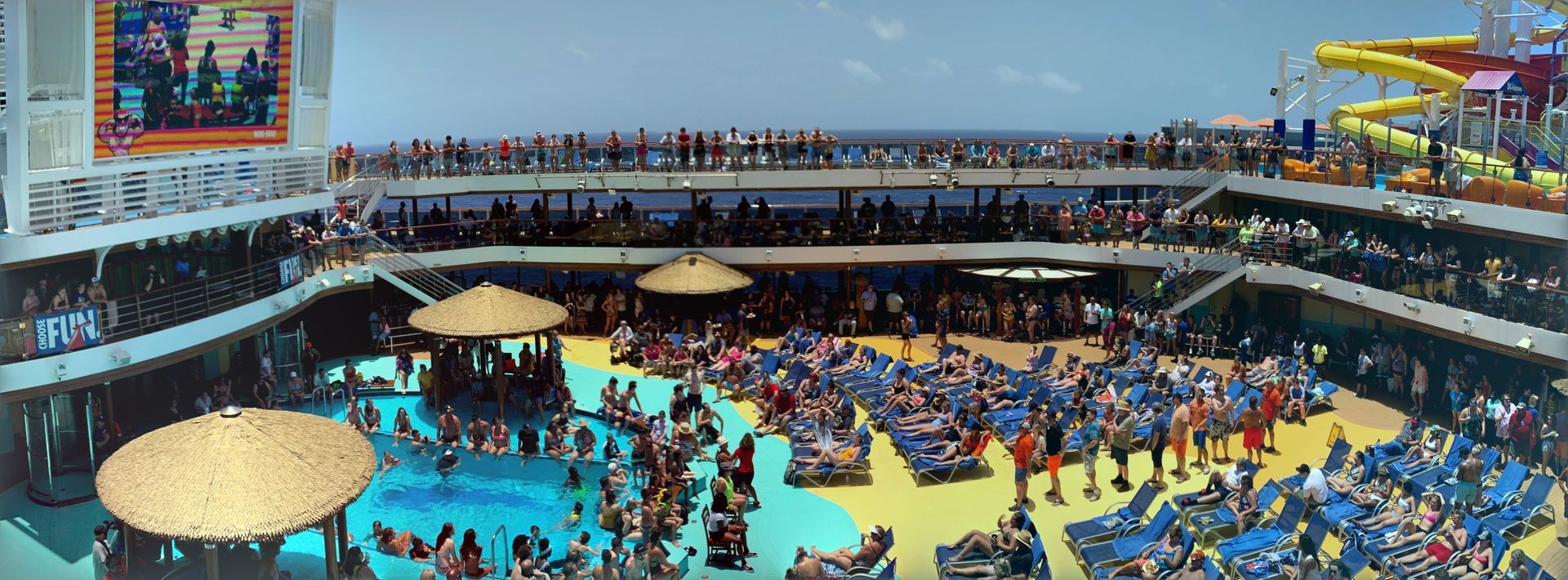 Crowded cruise ship pool deck with blue water, yellow lounge chairs, and many sunbathers under umbrellas