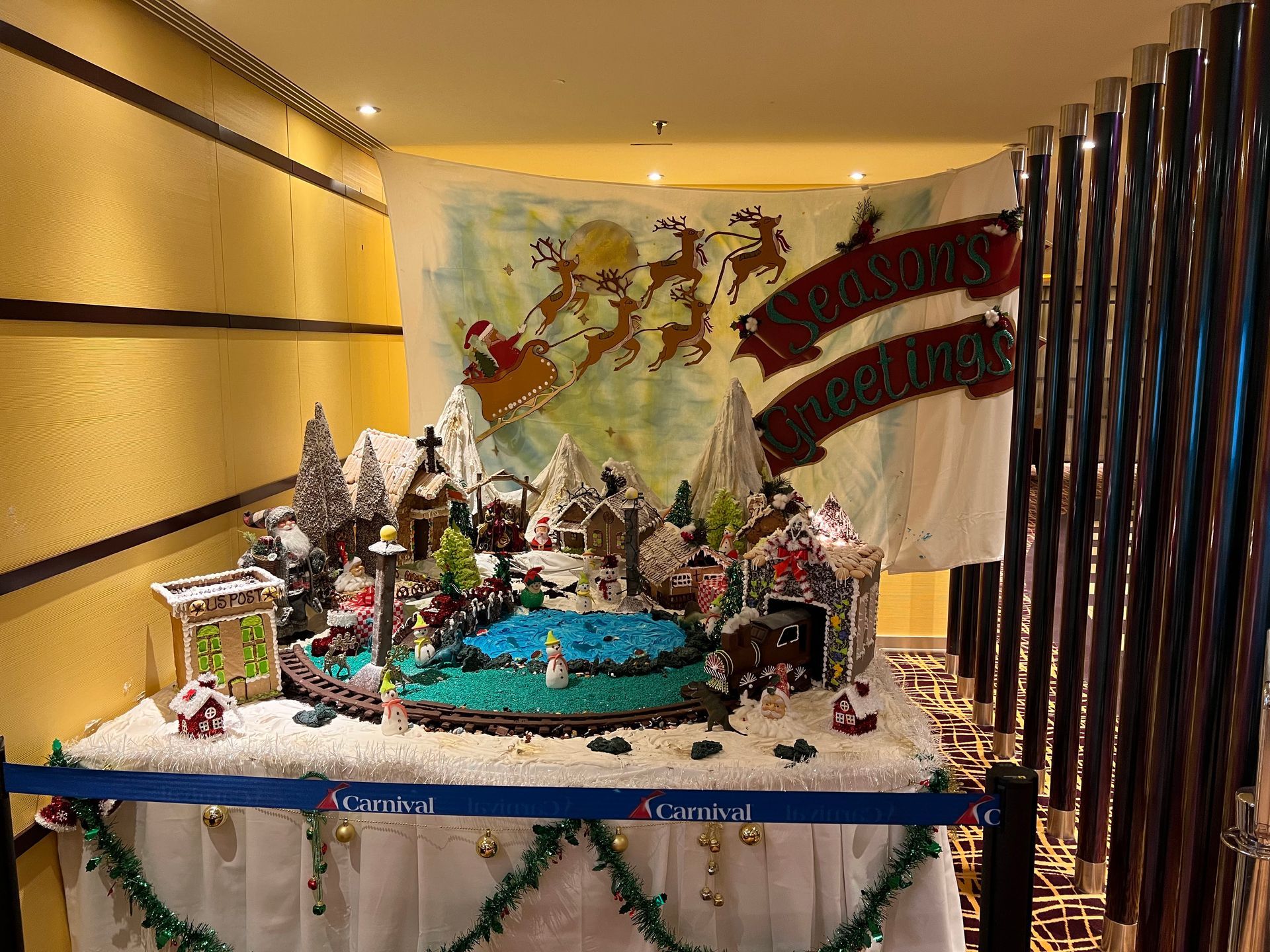Festive holiday display with a snowy miniature village, blue pond, and banner in a hallway