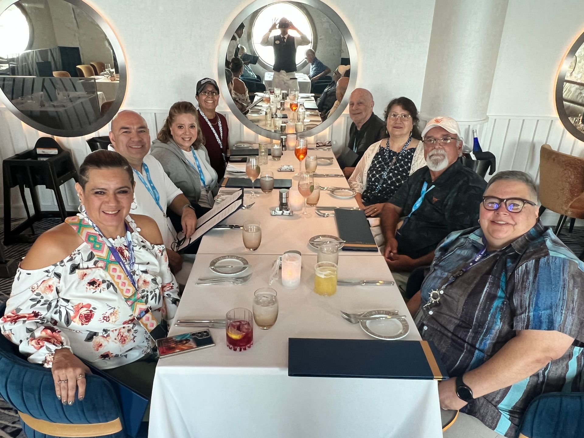 Group of people seated at a long dining table in a bright restaurant, with mirrored wall decor.