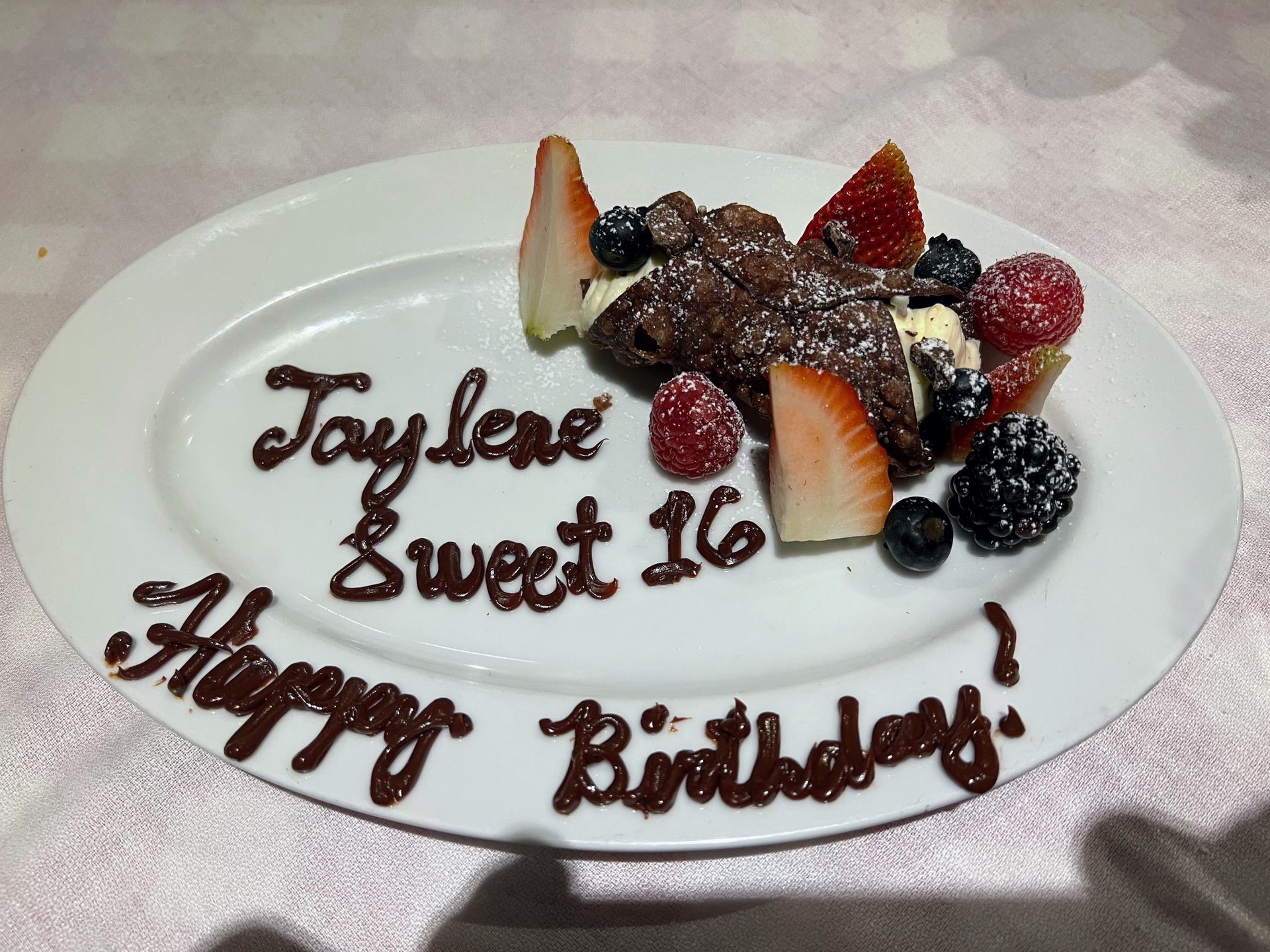 Birthday cake on white plate with chocolate writing: “Taylor Sweet 16 Happy Birthday,” topped with strawberries and berries