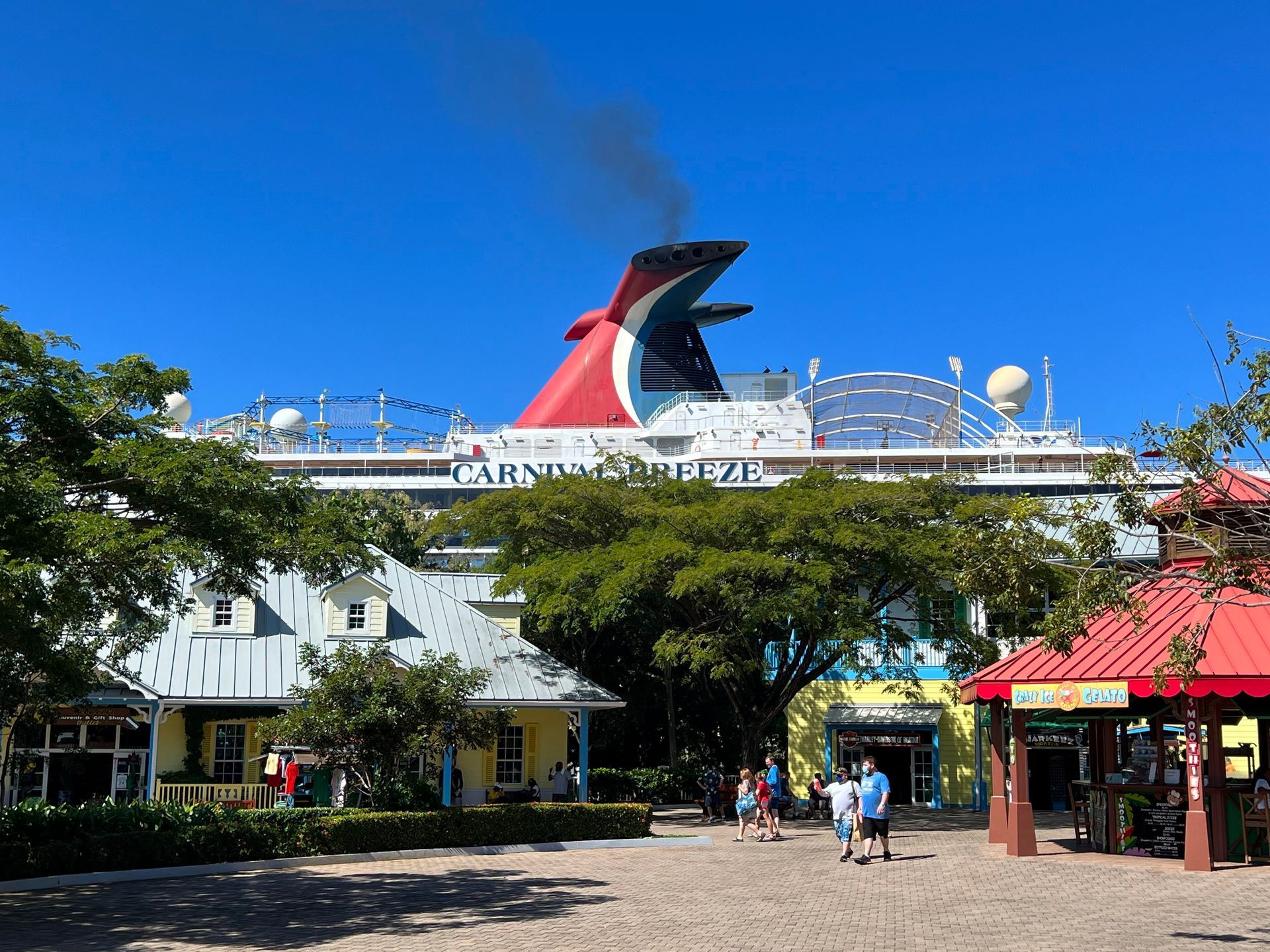Cruise ship deck entrance with red-and-black funnel, blue sky, and a few people walking below
