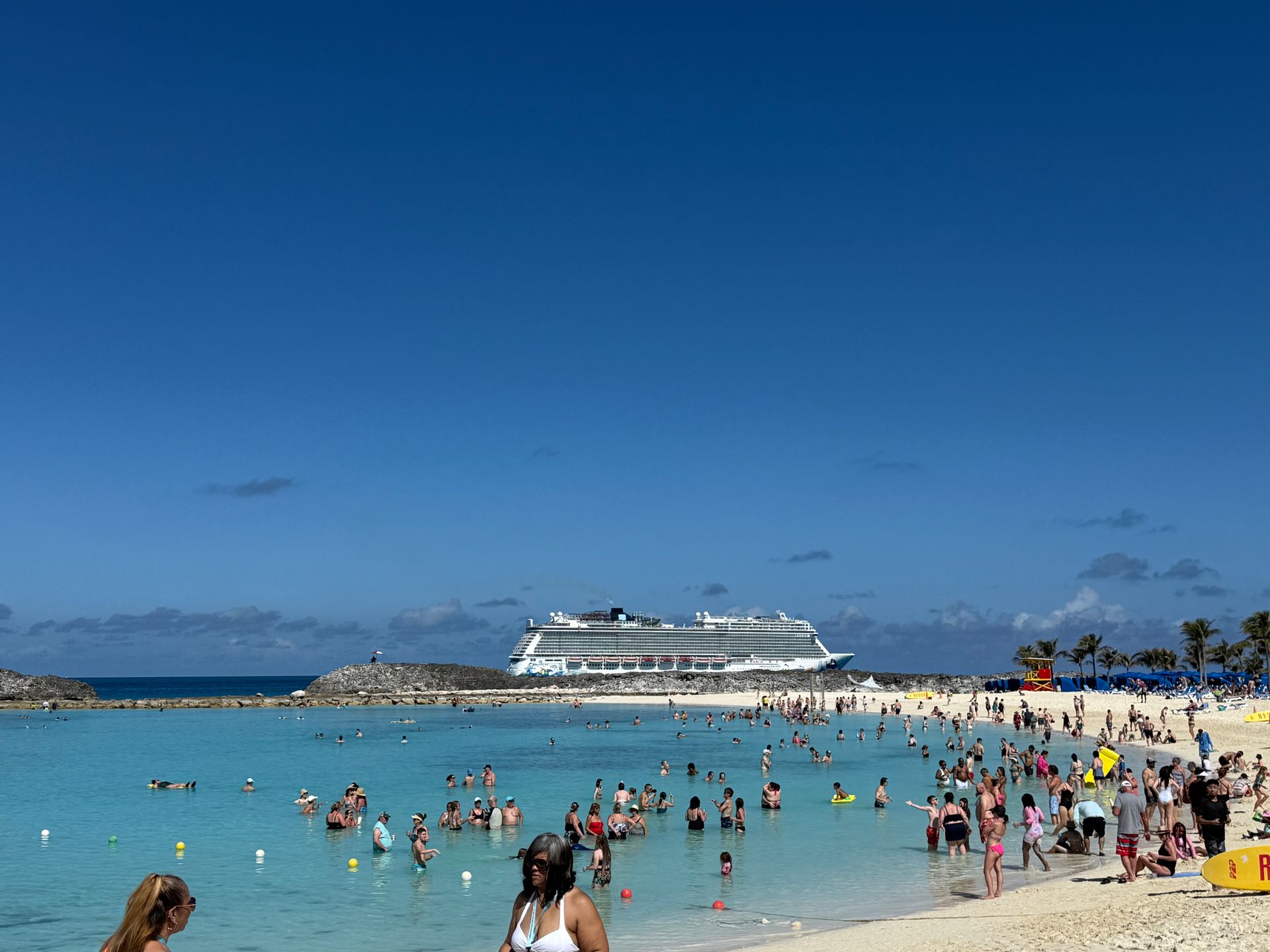 Crowded tropical beach with swimmers in turquoise water and a large cruise ship offshore