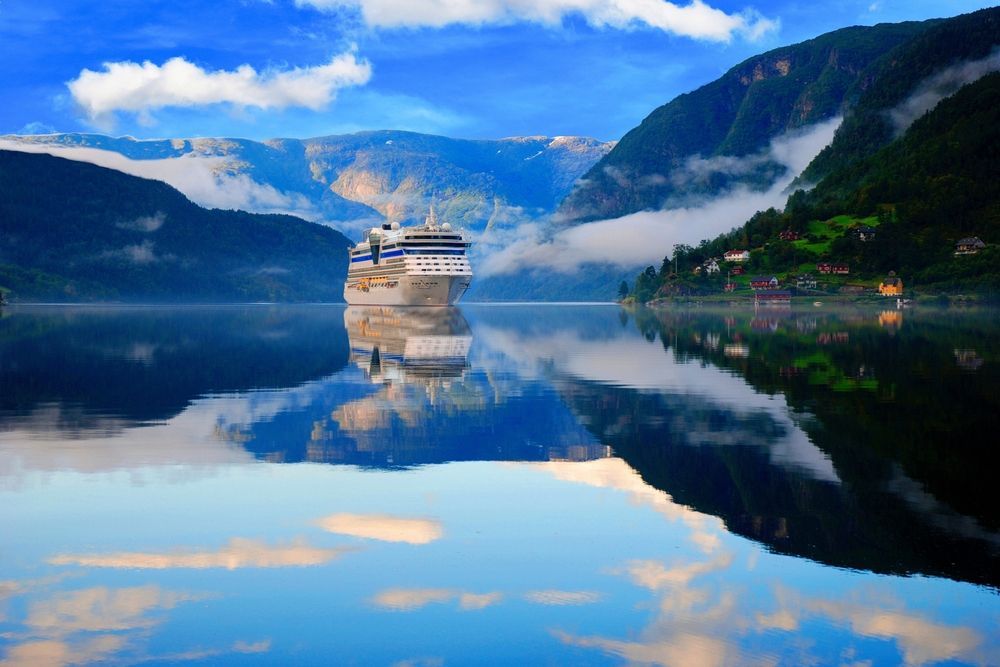 Cruise ship on a calm fjord, reflecting blue sky, mountains, and clouds at sunrise
