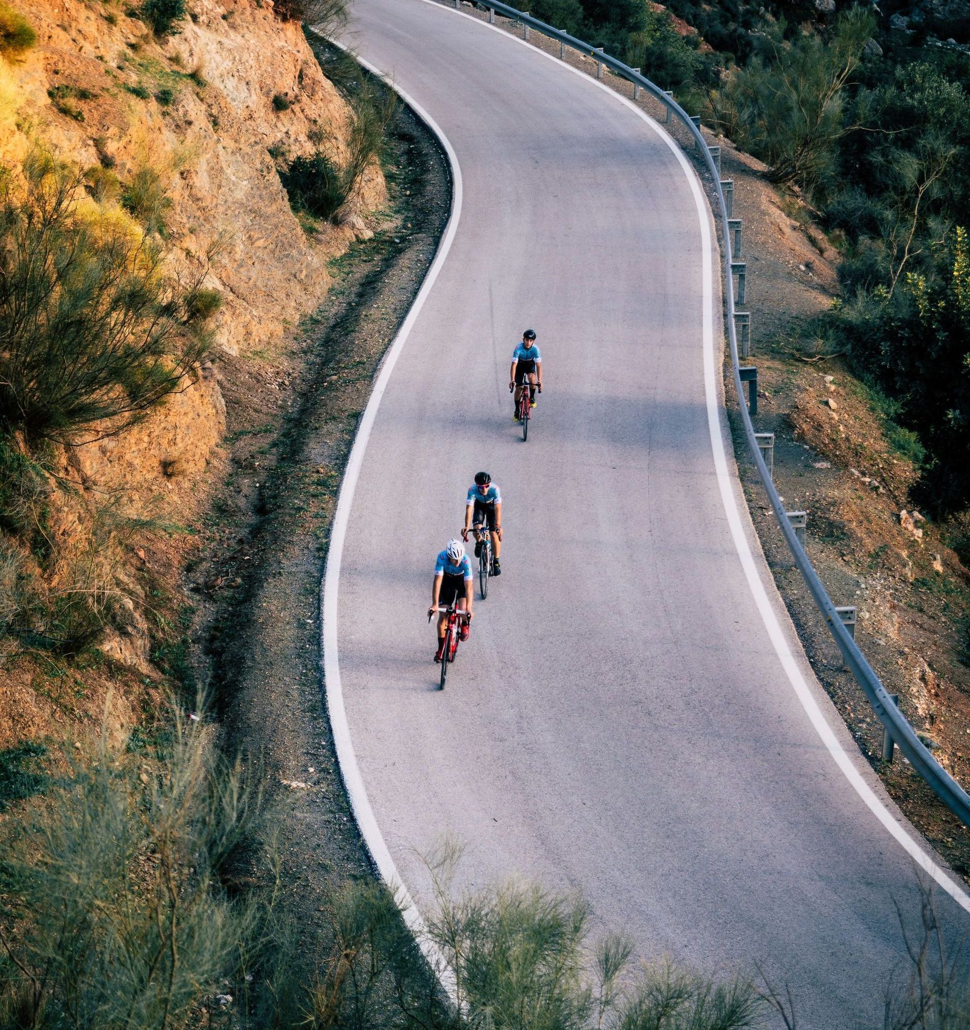 Three cyclists ride up a winding mountain road bordered by rocky slopes and greenery.