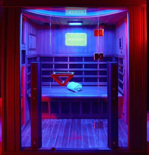 Neon-lit glass elevator interior with a bench, red accents, and a glowing sign behind it