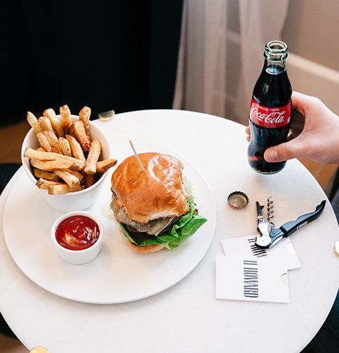 Burger, fries, and ketchup on a white table as a hand holds up a Coca-Cola bottle.