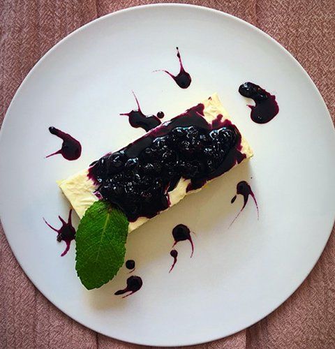 Dessert slice with dark berry topping and mint leaf on a white plate, garnished with sauce dots