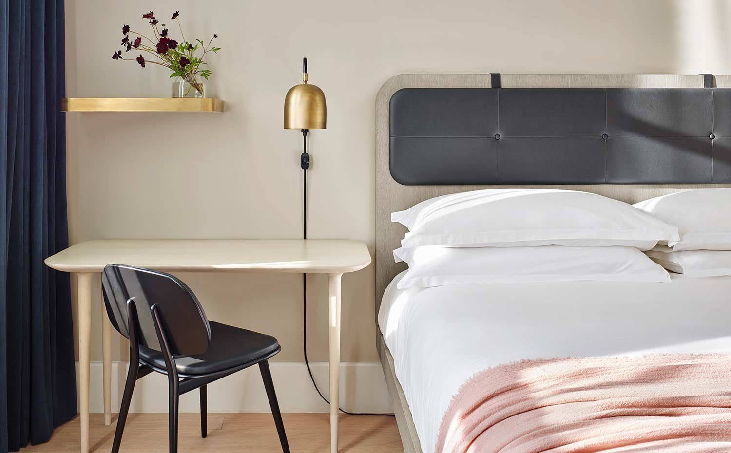 Modern bedroom with gray headboard, white bedding, small desk, black chair, gold lamp, and wall shelf with flowers