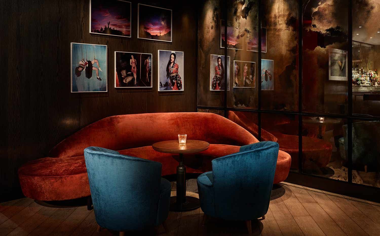A dimly lit lounge with a curved red velvet couch, two blue velvet chairs, and a small wooden table with a lit candle on it.
