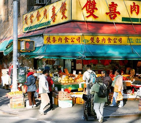 Busy street market storefront with yellow signs, teal awning, and shoppers browsing produce outside.