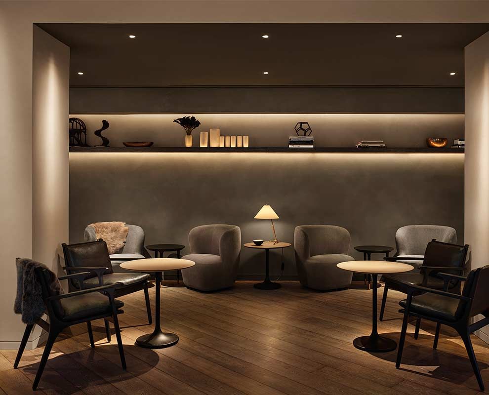 Dim modern lounge with armchairs, side tables, warm lighting, and wall shelves in a cozy seating area