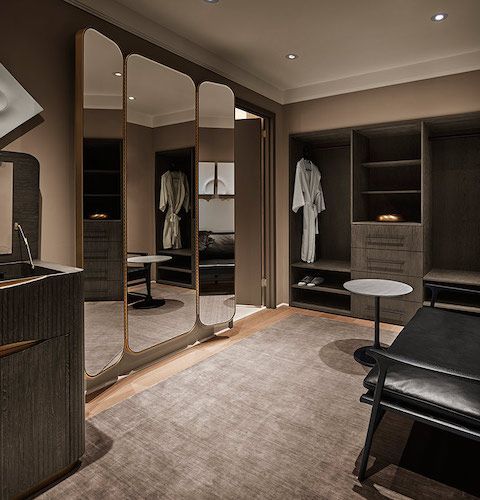 Modern walk-in closet with mirrored doors, open shelving, hanging clothes, and a chair on a neutral carpet.