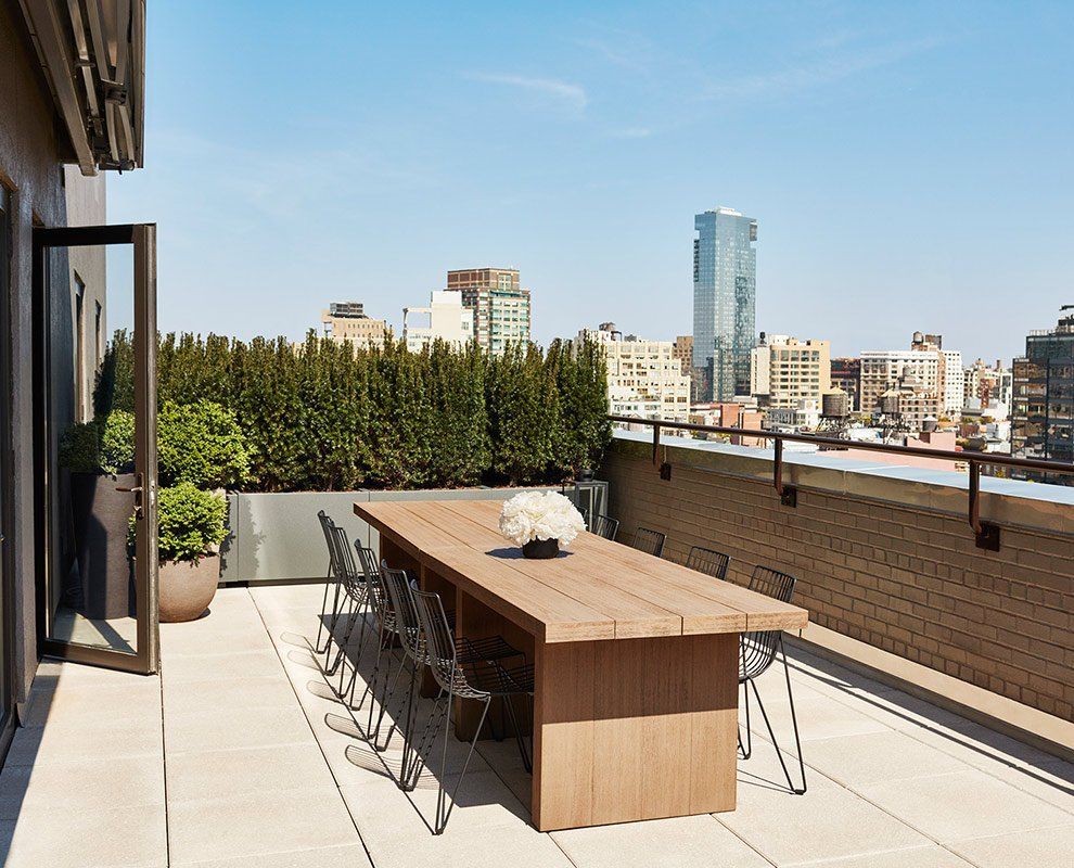 A rooftop terrace with a large wooden table and chairs, surrounded by plants and a city skyline.