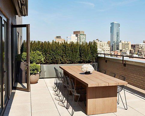 Modern rooftop terrace with wooden dining table, lounge chairs, potted plants, and city skyline view.