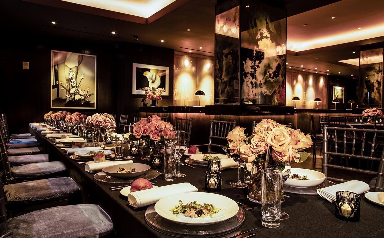 Elegant restaurant dining room with a long set table, pink floral centerpieces, and warm ambient lighting