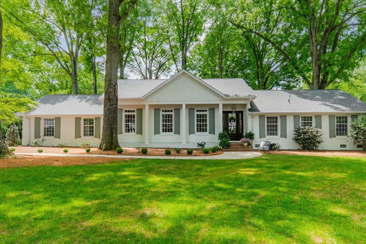 A white house with gray shutters and a large lawn in front of it surrounded by trees.