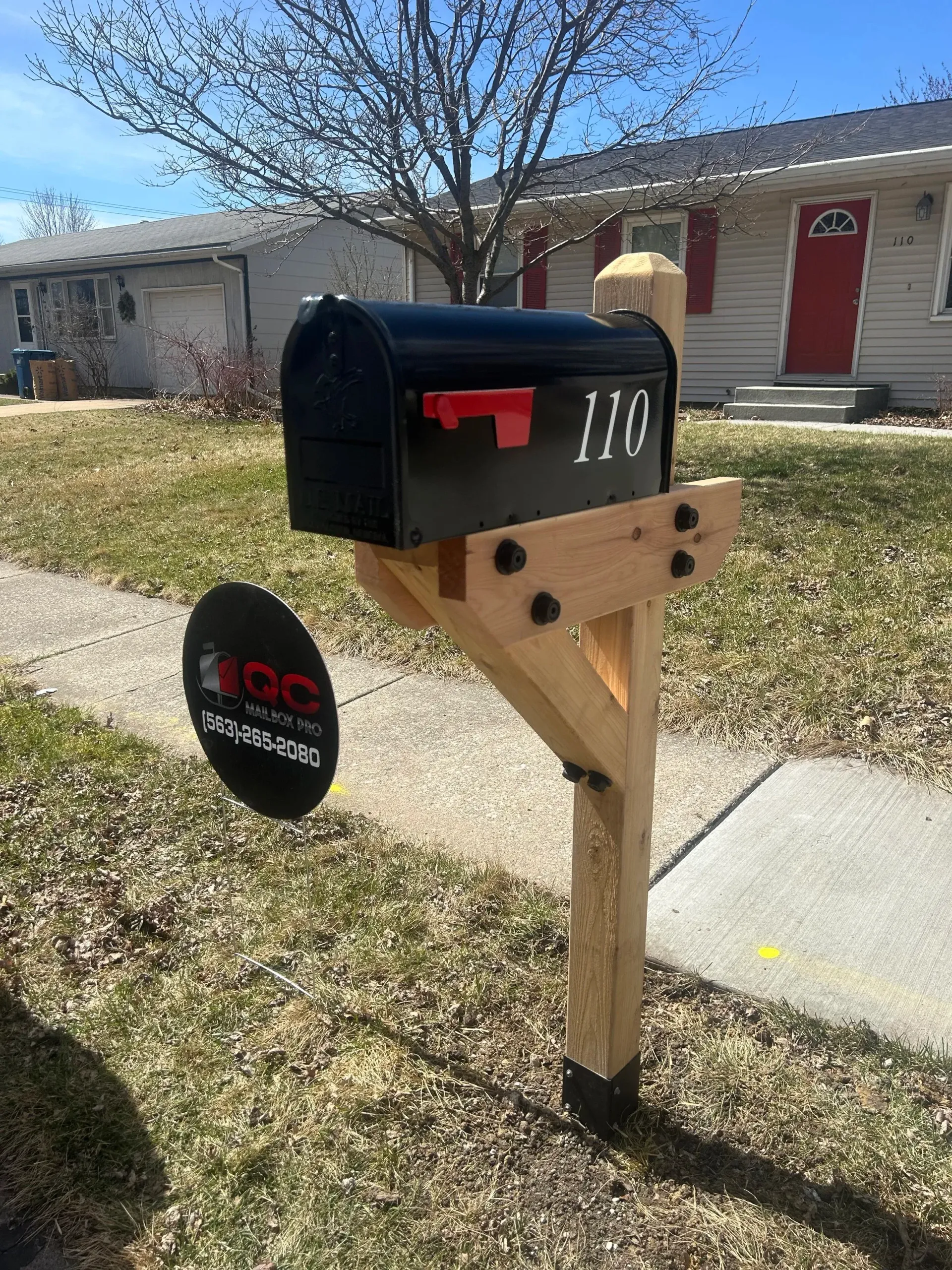 Black mailbox with red flag and house number 110 on a wooden post by a suburban sidewalk.
