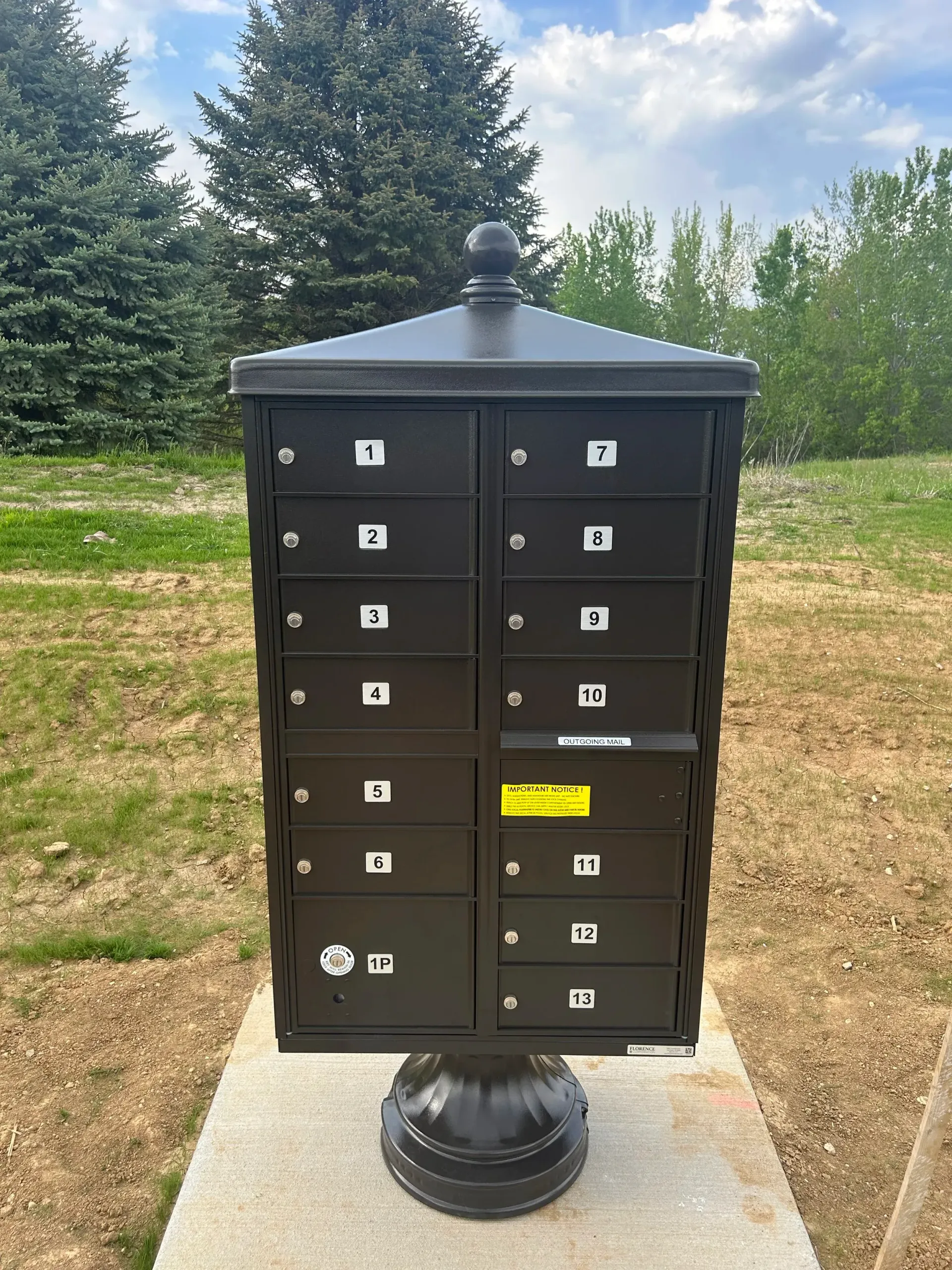 Black double mailbox stand with many slots, on a pedestal, outdoors near trees and grass