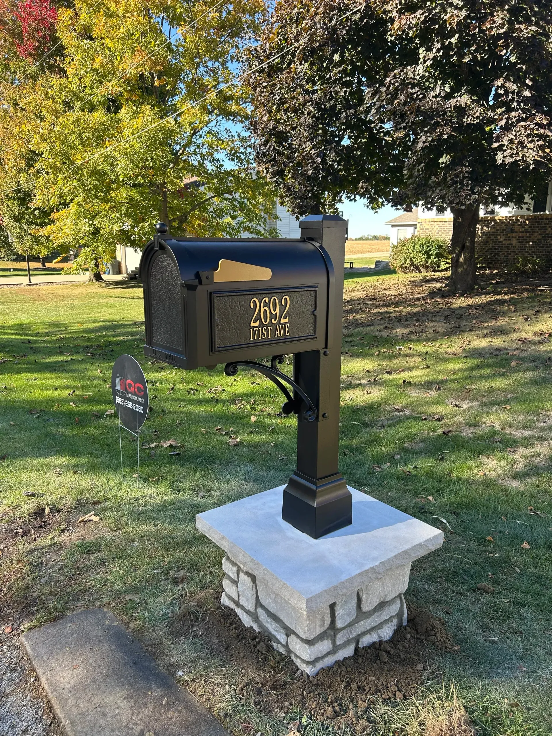 Black mailbox on a white stone pedestal in a grassy yard with trees in the background