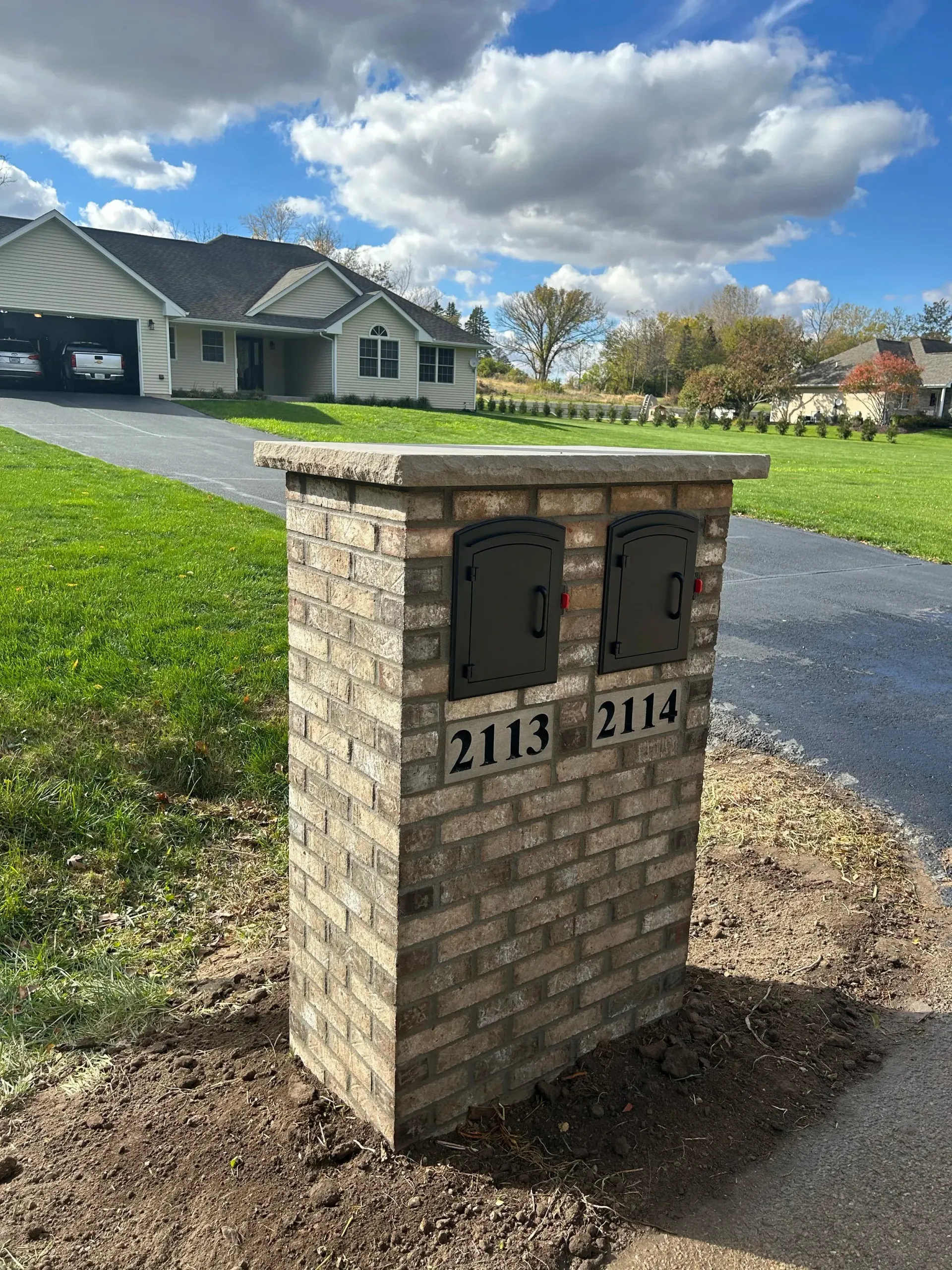 Brick mailbox post with two black mailboxes on a suburban street, house and cloudy sky in background