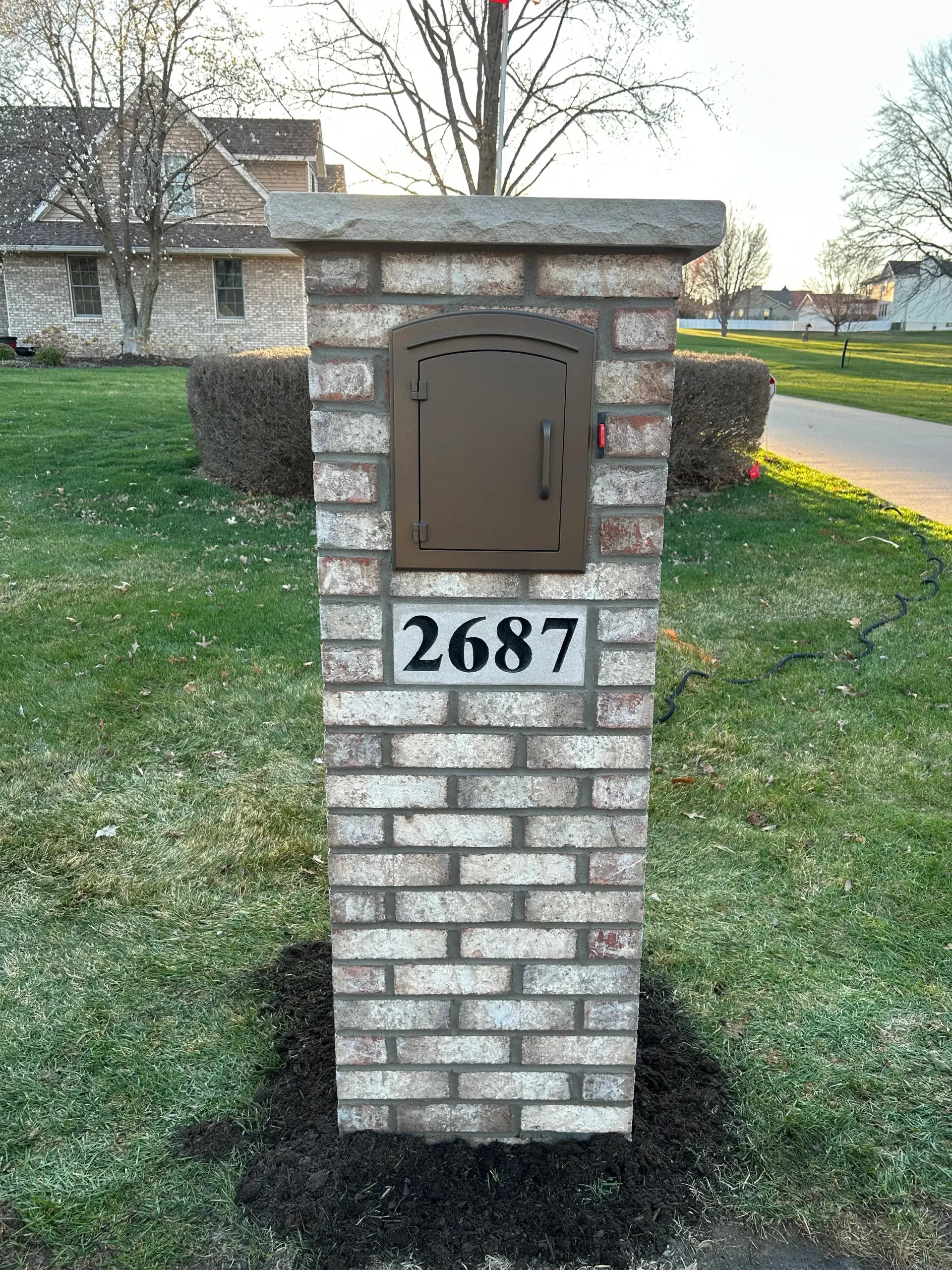 Brick mailbox pillar with brown mail slot and house number 2687 on a lawn near a road.