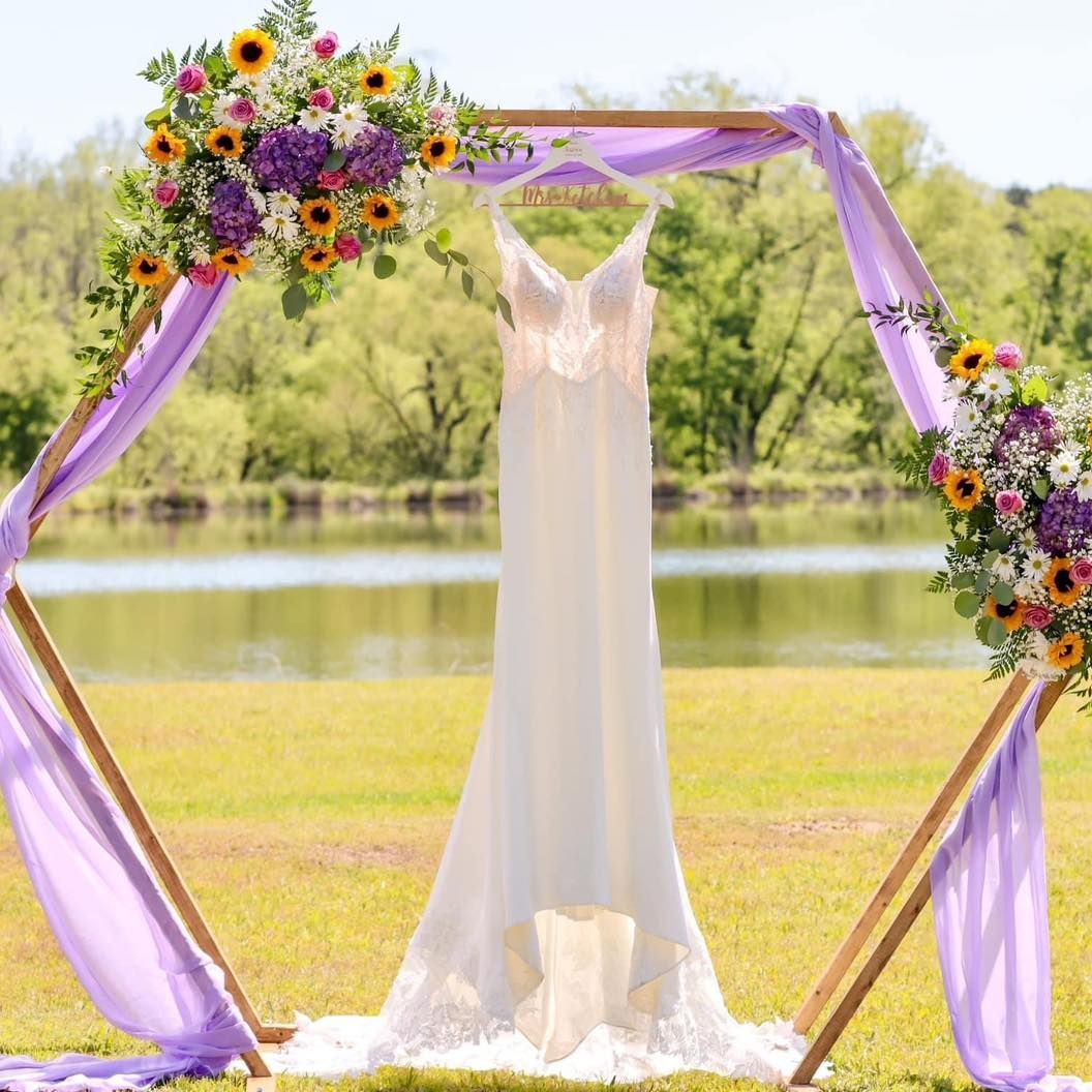 A wedding dress is hanging from a wooden arch decorated with flowers.