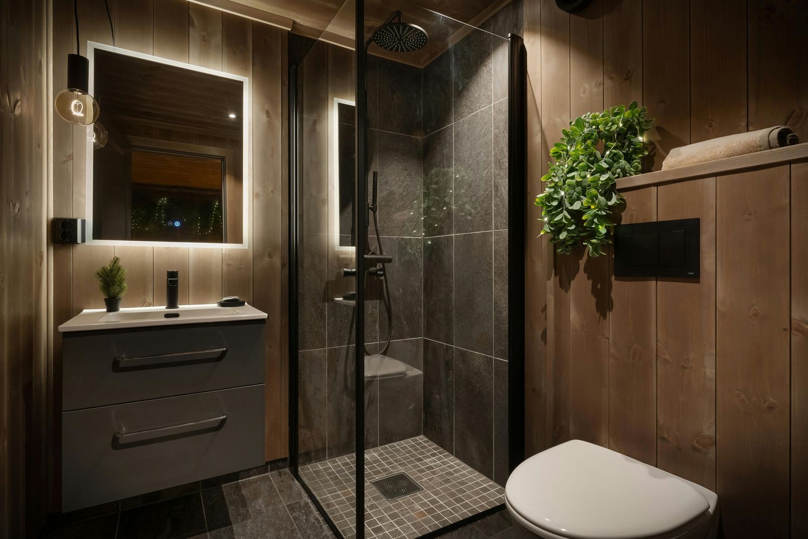 Modern bathroom with glass shower, vanity sink, wall mirror, and potted plant under warm lighting