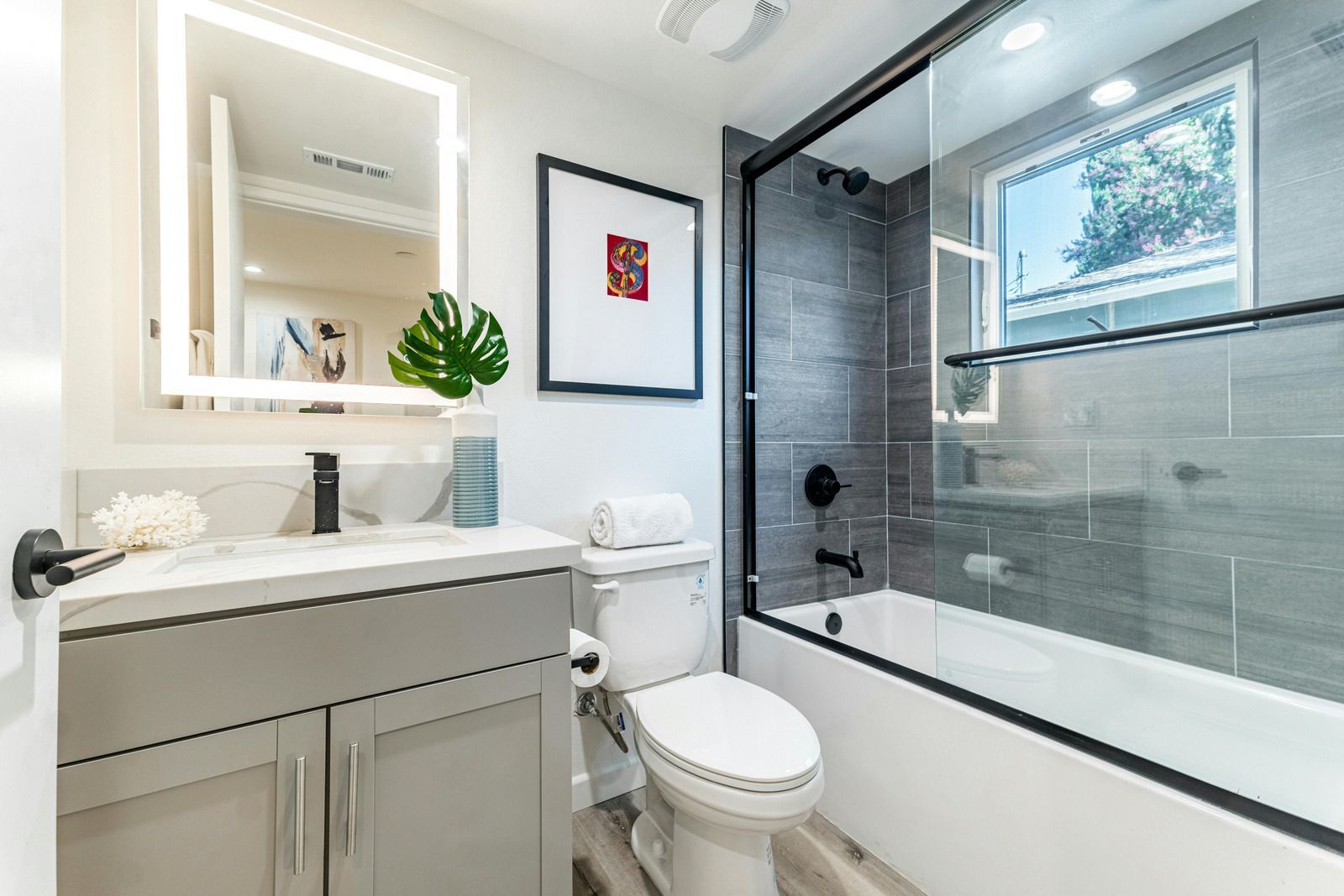 Modern bathroom with vanity, lit mirror, white toilet, and glass-enclosed shower with gray tile.