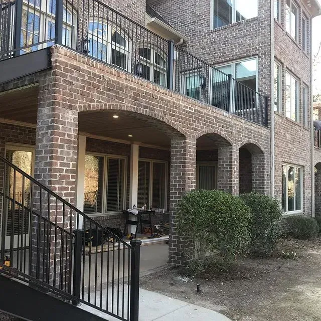 Brick apartment building with covered patio, black metal railing, and shrubs outside.