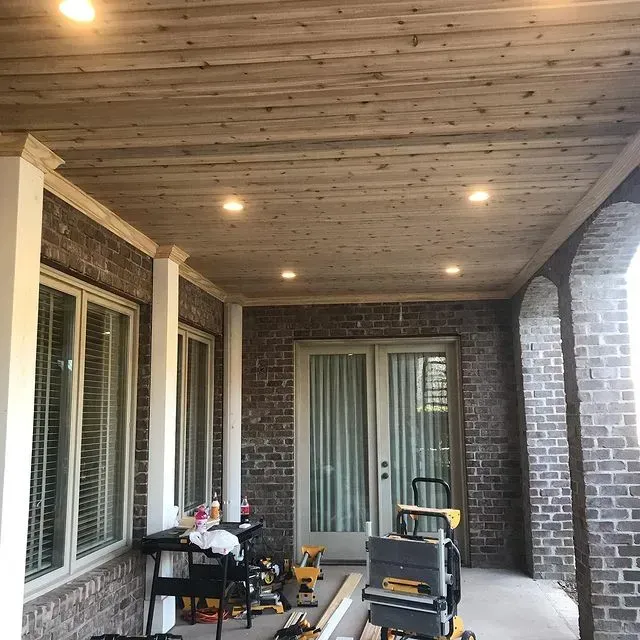 Covered brick porch with wood ceiling, recessed lights, windows, and a small work table and cart.