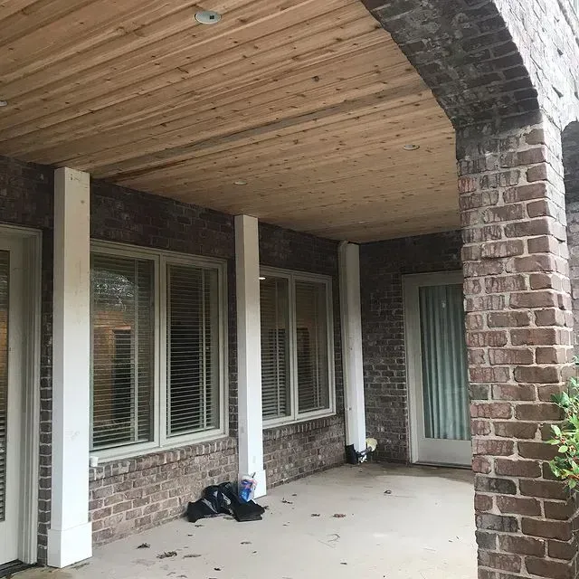 Covered brick porch with white columns, windows, and a glass door under a wood-paneled ceiling.