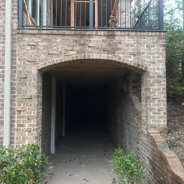 Brick archway leading into a dark underpass beneath a balcony, with brick walls and small shrubs.