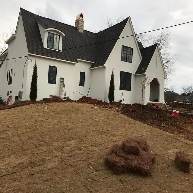 White house with dark roof on a sloped yard, with small shrubs and overcast sky.