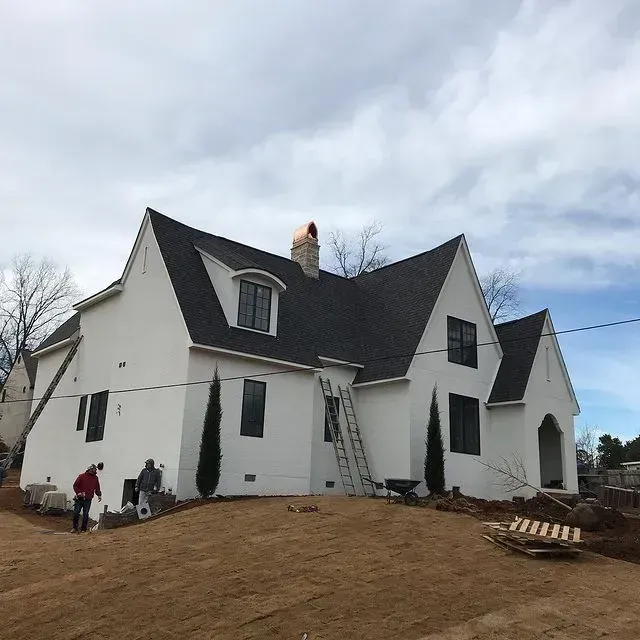 White house with dark roof under renovation, workers and ladders on a sloped dirt yard.