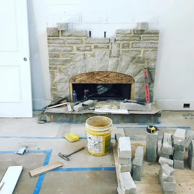 Stone fireplace under construction in a room, with tools, bucket, and stacked pavers on the floor.